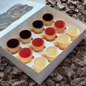 Könnte beinhalten: Eine offene Schachtel mit einem Dutzend essbaren Miniaturbechern. Die Becher haben eine hellbraune Außenseite und sind mit dunkler Schokolade, roter und weißer Schokolade gefüllt. Die Becher sind in Reihen angeordnet.