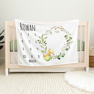 May include: White baby milestone blanket with the name "Rowan" printed at the top. The blanket features a watercolour wreath design with a dinosaur hatching from an egg. Numbers 1-12 are printed around the wreath, with the word "Months" at the bottom.