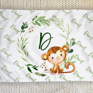 Könnte beinhalten: Weiße Babydecke mit einer Aquarellillustration eines Affen in einem grünen Blätterkranz. Der Buchstabe "D" befindet sich in der Mitte. Der Name "Daniel" ist in grüner Schrift über die Decke geschrieben.