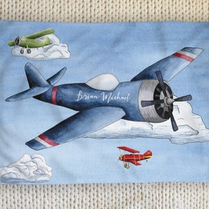 May include: Watercolor illustration of a blue airplane with red and white accents, flying in a light blue sky with clouds. The name "Brian Michael" is written on the plane. A green and red airplane are also depicted.