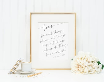 May include: A framed print with the word "love" and the phrase "bears all things, believes all things, hopes all things, endures all things, love never fails" in elegant script. A cup of coffee, pen, and flowers are also present.