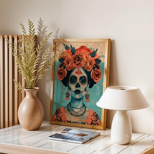 a framed artwork of a woman with a skull design, surrounded by flowers, placed on a table next to a lamp and a vase with greenery.
