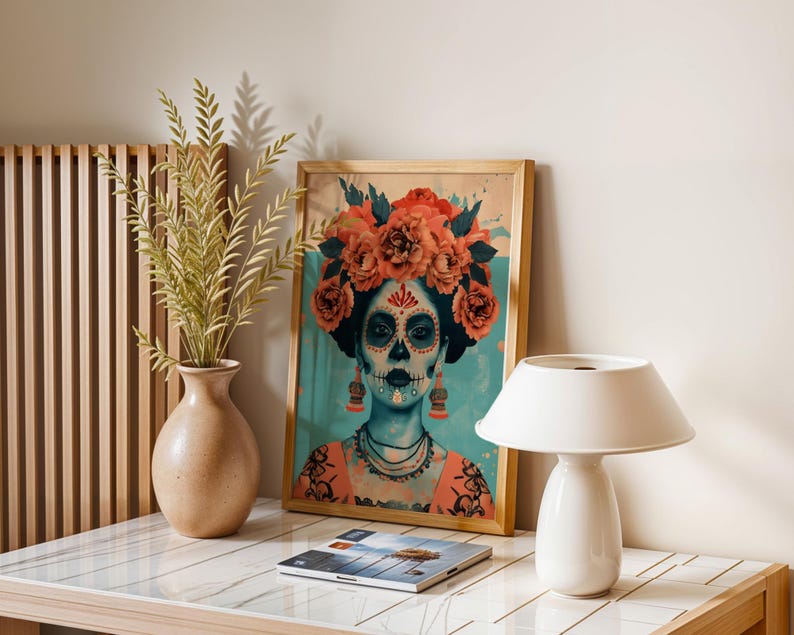 a framed artwork of a woman with a skull design, surrounded by flowers, placed on a table next to a lamp and a vase with greenery.