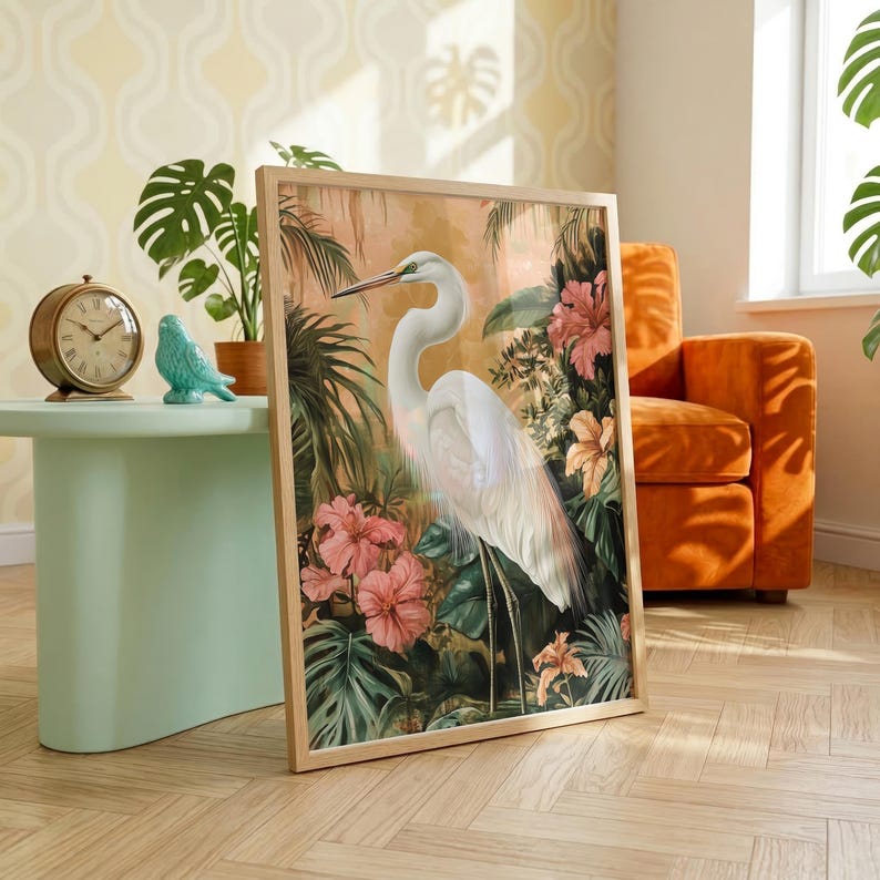a large framed painting of a white heron surrounded by tropical foliage, including pink flowers, in a room with a green table, a gold clock, and an orange armchair.