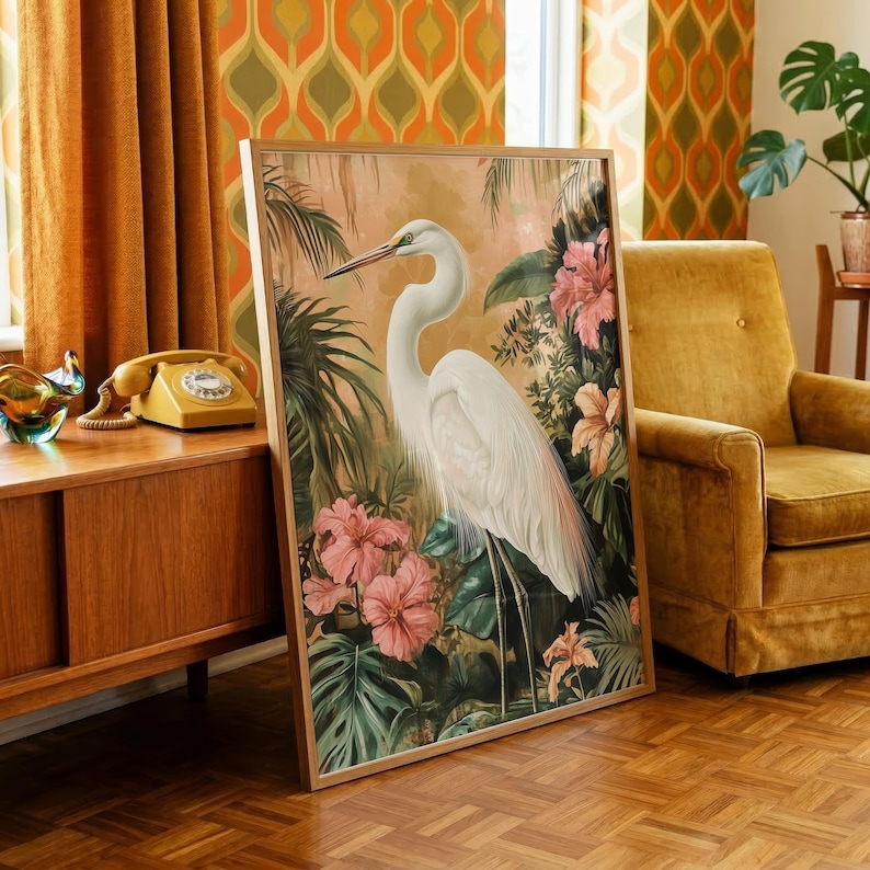 a large framed painting of a white heron surrounded by tropical foliage, including pink flowers, placed on a wooden floor in a room with a yellow armchair and a vintage telephone.