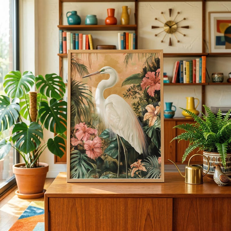a framed painting of a white crane standing amidst tropical foliage, including pink flowers, on a wooden surface.
