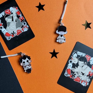 Peut inclure: Fond orange avec trois breloques de personnages de dessins animés et deux cartes noires. Les breloques ont des cordons blancs. Les cartes ont des illustrations et le mot "OFF". Des étoiles noires sont dispersées autour des objets.