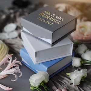 May include: A stack of four gift boxes, the top box is gray with gold lettering that reads "Mr. & Mrs. Burrell 11.11.2023". The boxes are stacked on a gray textured surface with white flowers.