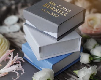 May include: A stack of four gift boxes, the top box is gray with gold lettering that reads "Mr. & Mrs. Burrell 11.11.2023". The boxes are stacked on a gray textured surface with white flowers.