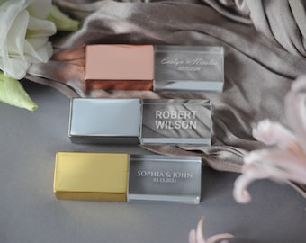 May include: Three crystal USB drives with gold, silver, and rose gold metal accents. The drives are engraved with names and dates.  The drives are on a gray surface with a white flower and a pink flower.