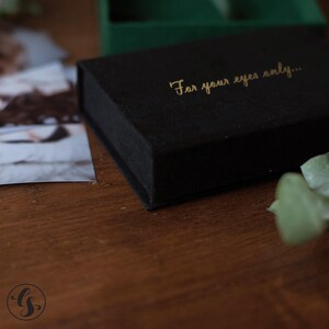 May include: A black velvet box with gold lettering that reads "For your eyes only...". The box is rectangular and sits on a wooden surface. A photograph is partially visible to the left of the box.