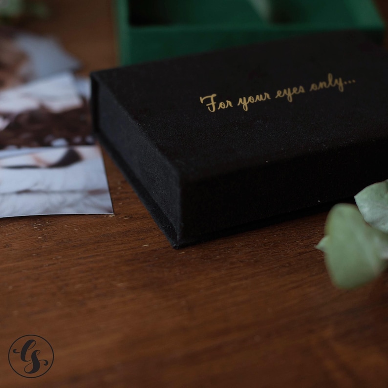 May include: A black velvet box with gold lettering that reads "For your eyes only...". The box is rectangular and sits on a wooden surface. A photograph is partially visible to the left of the box.