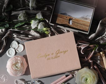 May include: A blush-colored wedding photo box with gold lettering that reads "Evelyn & George 03.15.2024" is surrounded by white flowers, wax seals, and a photo album. The album displays a wedding photo.