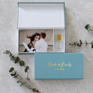 May include: A teal rectangular presentation box with a hinged lid. The lid features gold foil text reading Noah and Emily with a heart icon and the date 6.16.2026. Inside, a photograph of a couple and a small gold-topped clear glass vial are stored.