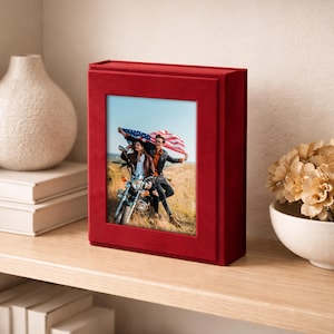 May include: A red velvet photo album with a rectangular frame. The album displays a photograph of a couple on a motorcycle holding an American flag. The album is on a wooden shelf, next to a white vase and a small floral arrangement.