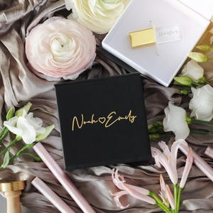 May include: A black box with gold lettering that reads "Noah & Emily" sits on a draped fabric surface, surrounded by white and pink flowers. A gold USB drive is in an open white box. Pink wax sticks and a gold wax stamp are also present.