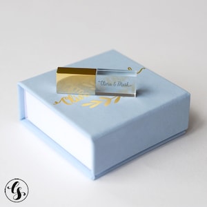 May include: A gold and clear USB flash drive with the names "Olivia & Mark" printed on it, resting on a light blue box. The box has a white interior and gold leaf design. The USB drive has a gold cap.