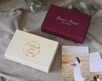 May include: Two rectangular boxes, one ivory and one burgundy, with gold and silver text. The ivory box has "Matthew and Claire" in a wreath. The burgundy box reads "Janet & Patrick 03.15.2023". Wedding photos are also visible.