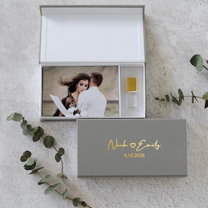 May include: A gray rectangular keepsake box with a hinged lid, featuring gold foil text that reads Noah Emily and the date 6.16.2026. Inside, a photograph of a couple is displayed alongside a small, clear glass vial with a gold cap.