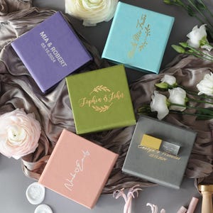 May include: An assortment of colorful, square gift boxes with gold lettering and dates. The boxes are in shades of purple, green, pink, blue, and gray. The boxes are arranged on a satin fabric with flowers.