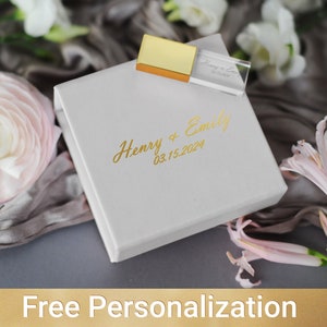 May include: A white box with a gold inscription that reads "Henry & Emily 03.15.2024". A clear crystal USB drive with a gold top is sitting on top of the box.