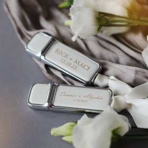 May include: Two white USB drives with silver trim. The drives are personalized with the names of couples and their wedding dates. The text on the drives reads "Rick + Maci 11.18.23" and "Conner + Alyshia 8.10.2023".
