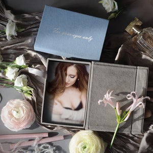 May include: A gray velvet photo album with a blue velvet lid. The lid has the text "For your eyes only" written in white. The album is open and a photo of a woman is visible inside. The album is surrounded by flowers and other decorative items.