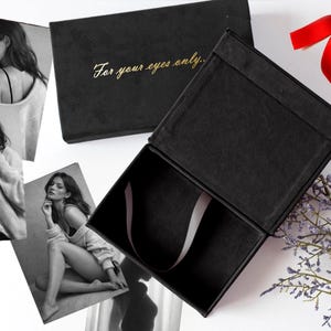 May include: Black gift box with the text "For your eyes only" in gold lettering. The box is open, revealing a satin ribbon. The image also includes black and white photographs of a woman, red heart decorations, and a red ribbon.