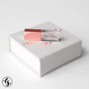 May include: A rose gold USB drive with the text "Olivia & Mark" on a white box with rose gold floral accents. The box is square and has a hinged lid. The USB drive is rectangular with a clear plastic body.