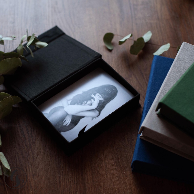May include: A black photo box with a black and white photograph inside. The photograph depicts a person with long hair. Beside the box are three hardcover books in blue, beige, and green. Eucalyptus leaves are in the background.