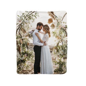 Puede incluir: Una pareja con atuendo de boda se encuentra bajo un arco floral en una playa. La novia lleva un vestido blanco y el novio lleva una camisa blanca y pantalones negros. El arco está decorado con flores blancas y vegetación.