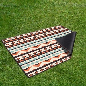 Puede incluir: Alfombra rectangular para exteriores con un patrón geométrico en crema, marrón, turquesa y naranja. La alfombra está sobre hierba verde, con una esquina levantada, revelando un reverso negro. El diseño presenta diamantes y zigzags.