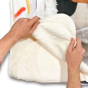 Op de afbeelding: Een wit sherpa fleece deken met een bedrukt ontwerp aan de ene kant. Het deken wordt vastgehouden door de handen van een persoon, waardoor de zachte, pluche textuur van de sherpa zichtbaar wordt.