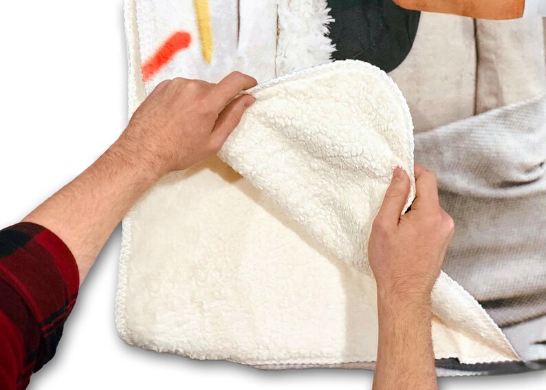 Op de afbeelding: Een wit sherpa fleece deken met een bedrukt ontwerp aan de ene kant. Het deken wordt vastgehouden door de handen van een persoon, waardoor de zachte, pluche textuur van de sherpa zichtbaar wordt.