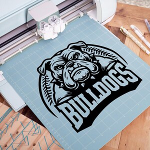 May include: A black and white graphic design of a bulldog's head with the word "Bulldogs" in a bold, black font. The design is on a blue cutting mat.