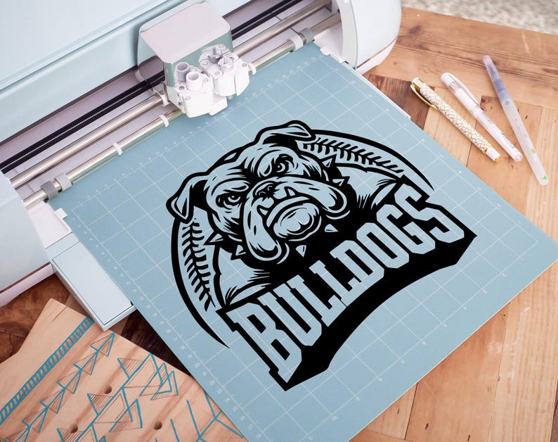 May include: A black and white graphic design of a bulldog's head with the word "Bulldogs" in a bold, black font. The design is on a blue cutting mat.