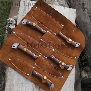 May include: A set of five knives with wooden handles and leather sheaths, rolled up in a brown leather carrying case. The case has the text "Metal Layers" printed on it.