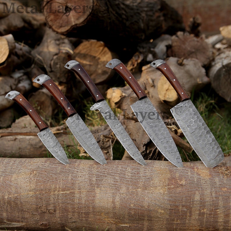 May include: Set of six kitchen knives with wooden handles and patterned blades. The knives are arranged on a wooden log.