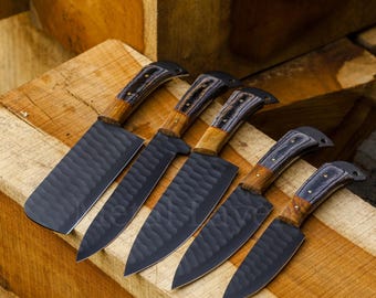 May include: Five black kitchen knives with wooden handles and black and brown patterned inlays. The knives are arranged on a wooden surface.