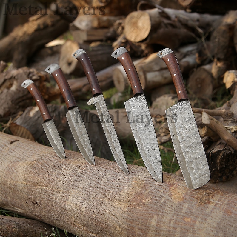 May include: Set of six kitchen knives with wooden handles and patterned metal blades. The knives are arranged on a wooden log.