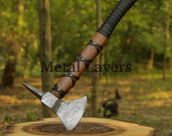 May include: A hand-forged axe with a wooden handle wrapped in black leather and brass accents. The axe head has a Damascus steel pattern and a pointed tip.