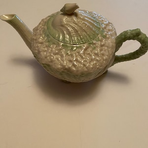 Könnte beinhalten: Eine Keramik-Teekanne mit Muscheldesign. Die Teekanne ist überwiegend cremefarben mit grünen Akzenten auf Deckel und Griff. Ausguss und Griff sind gebogen. Der Deckel hat einen kleinen knauf in Muschelform.