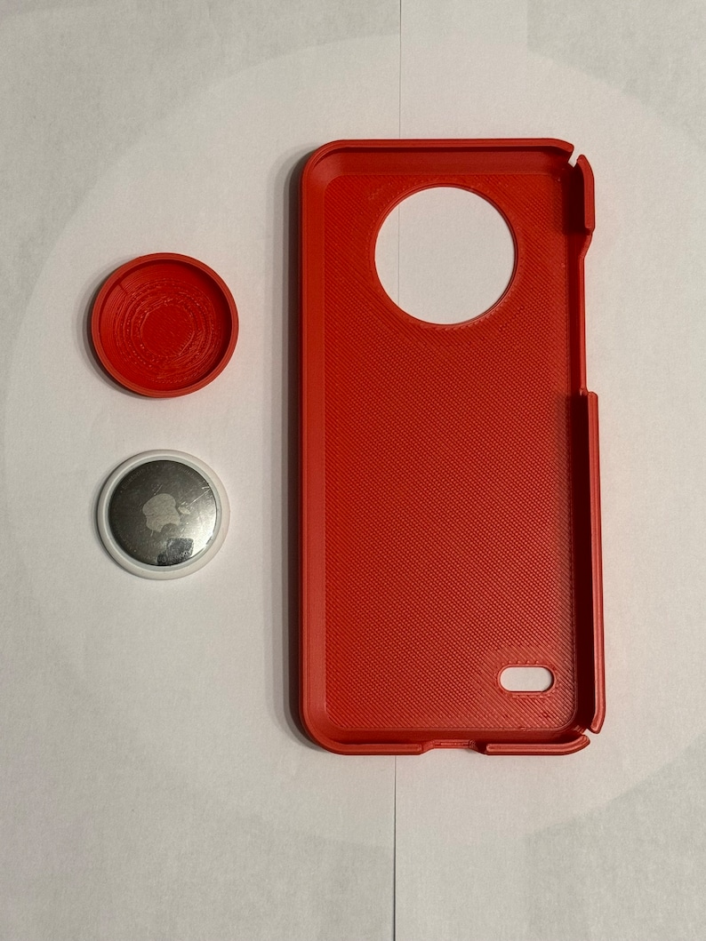 May include: A red phone case with a large circular cutout on the back. The case is made of a textured material. There are two small round objects next to the case. One is red and the other is white with a silver apple logo.