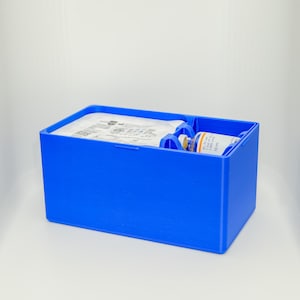 Pode incluir: Um recipiente de armazenamento de plástico azul brilhante com divisórias internas. Contém um pacote branco e um pequeno frasco com a etiqueta "SC IV use 1,000 units of the 10 ml". O recipiente está sobre uma superfície branca.