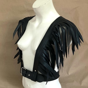 Peut inclure: Un gilet en cuir noir avec des détails en franges. Le gilet a un col en V et une ceinture avec une boucle argentée.