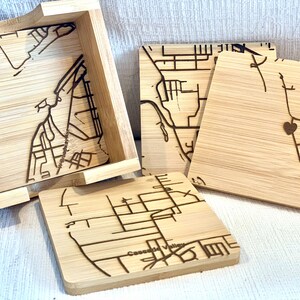 May include: Set of four bamboo coasters with a matching holder. Each coaster features an engraved map design with street details. The coasters are light brown with dark brown engraved lines. The holder is a small wooden box.