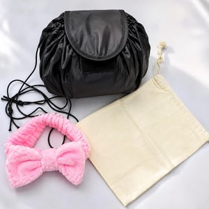 May include: A black drawstring cosmetic bag with a flap closure, a soft pink plush headband featuring a large bow detail, and a plain beige fabric drawstring pouch. The black bag has thin black cords and a structured, rounded shape.