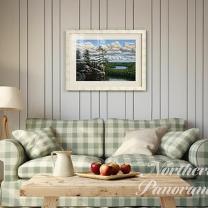 May include: A framed landscape painting of a lake and forest scene with a white frame and a white mat. The painting is hanging on a white wood wall in a living room setting with a green and white checkered couch and a wooden coffee table.