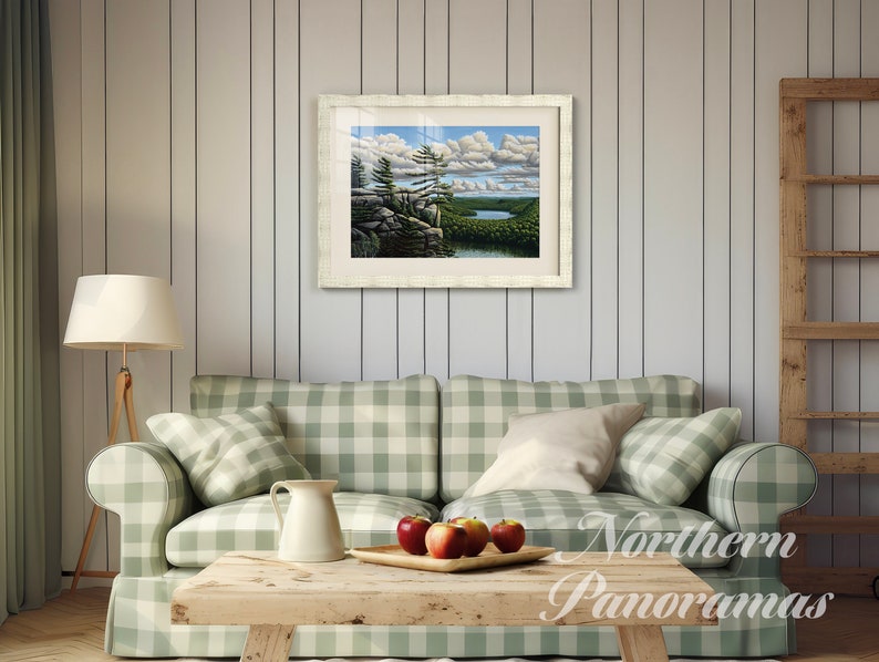 May include: A framed landscape painting of a lake and forest scene with a white frame and a white mat. The painting is hanging on a white wood wall in a living room setting with a green and white checkered couch and a wooden coffee table.