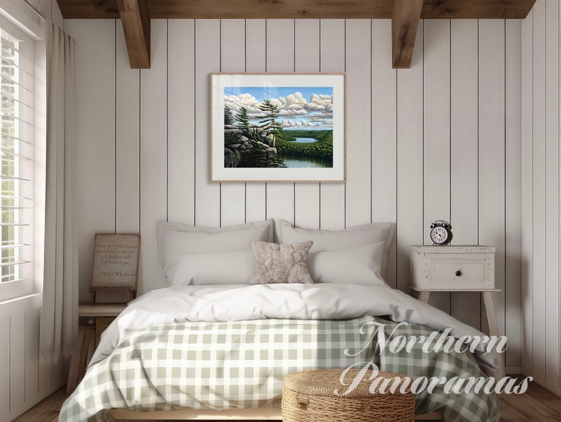 May include: A framed landscape painting of a lake and forest scene with a blue sky and white clouds. The painting is hanging on a white wood paneled wall in a bedroom with a bed, nightstand, and lamp.